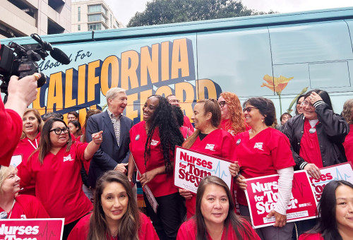 Tom Steyer (back left, in dark suit), the billionaire climate activist running for California governor, pauses for photos with members of the California Nurses Association, a progressive union that endorsed him in part due to his strong support of single-payer healthcare. (Christine Mai-Duc/KFF Health News)